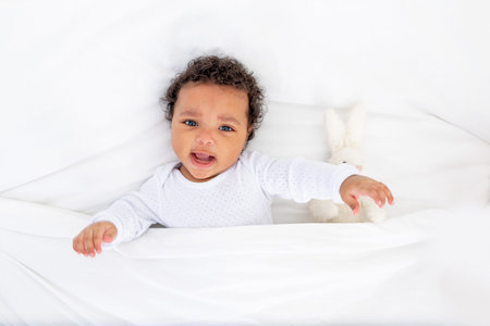 Smiling African-American little baby under a blanket with a toy on a white bed for sleeping