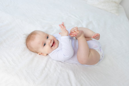 smiling baby girl six months old plays with her legs lying on her back on a white cotton bed in the...