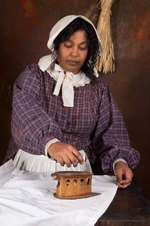 Reenactment scene of a black victorian maid ironing with an antique ironの写真素材