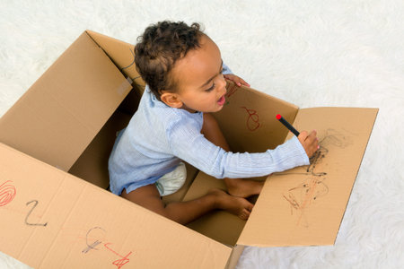 Mixed race little toddler boy sitting in a cardboard box playing with crayonsの写真素材
