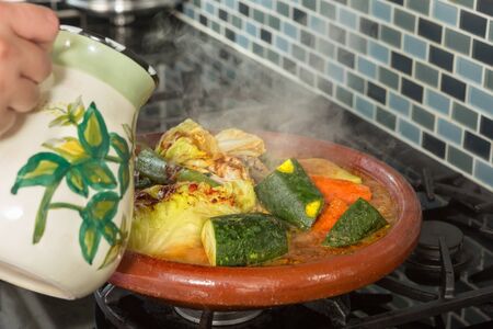 Hands of a woman adding water to a traditional Moroccan tajine during Ramadan nights (Moroccan immigrant woman in modern European kitchen)の写真素材