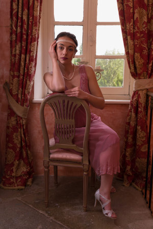 Pretty young woman in pink 1920s Gatsby flapper dress and headband sitting in antique interior