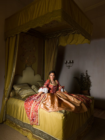 Golden medieval bedroom with beautiful canopy bed.  A lady in renaissance dress is sitting on the bed.の写真素材
