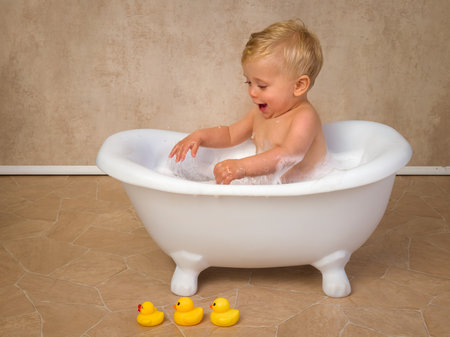 Happy scenes of a little cute baby boy of 12 months old sitting in a vintage white bath tub splashing with water and soapの写真素材