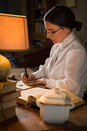 Beautiful lady in Edwardian skirt and high collar lace blouse sitting in the library. She could be a teacher or even a headmistress.の写真素材