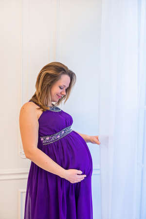 Beautiful pregnant woman in ultra violet dress standing near windowの写真素材