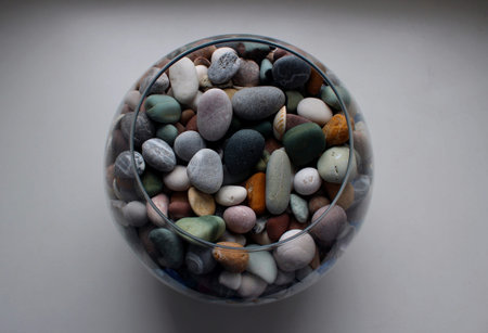 Round glass vase with colorful smooth pebbles and shells on a white surface isolated top view