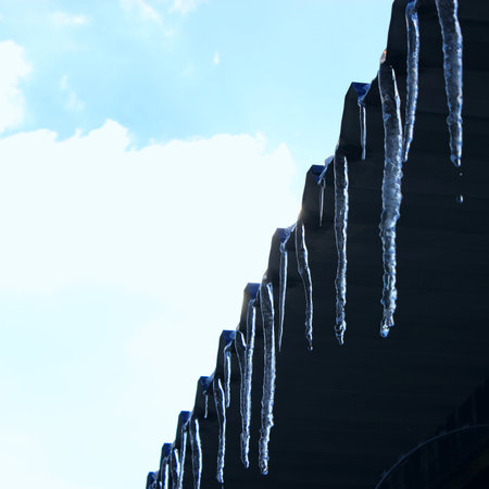 Melting transparent icicles on a metal roof eaves against the background of a spring blue sky with...