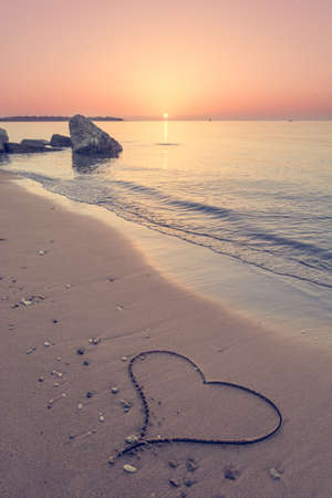 Heart drawn on the beach sand with sun above the horizonの写真素材