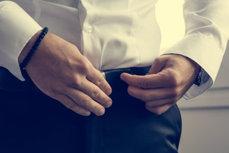 Groom preparing for wedding. Closeup of hands tying a belt.