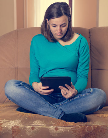Pregnant woman reading a tablet on a couch.