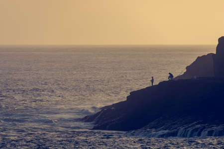 Pair of fisherman fishing from coastline at sunset.の写真素材