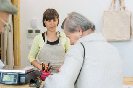 Young shopkeeper interacting with elderly lady at cash desk.の写真素材