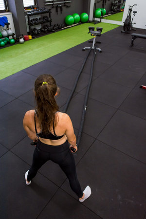 Young athletic woman exercising in gym using battle rope.