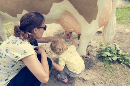 Cute blonde girl learning how to milk a cow on milking simulator with her mother.の写真素材