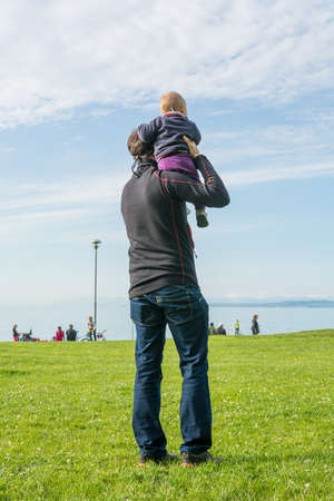 Rear view of a father holding his cute baby daughter and looking the sea.の写真素材