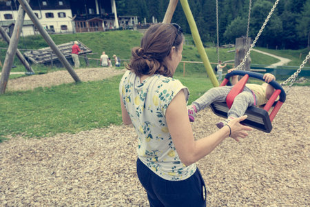 Mother pushing her daughter on a swing at outdoor playground. Happy and active parenthood.