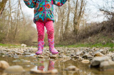 Young girl walking along a shallow stream with rubber pink boots. Long angle view of happy child.