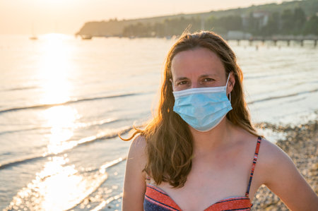 Young woman wearing medical mask at the beach during sunset.