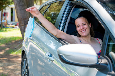Woman stretching her arm through a car window while holding a medical mask.
