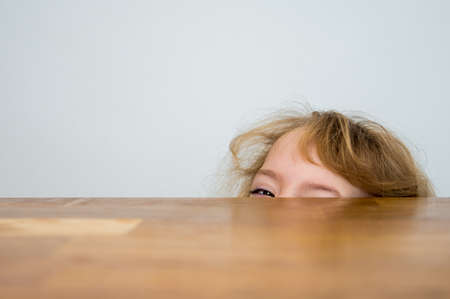 Cute blonde girl hiding half of her face under the wooden table.の写真素材