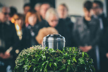 A metal urn with ashes of a dead person on a funeral, with people mourning in the background on a memorial service. Sad grieving moment at the end of a life. Last farewell to a person in an urn.の写真素材