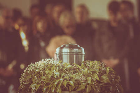 A metal urn with ashes of a dead person on a funeral, with people mourning in the background on a memorial service. Sad grieving moment at the end of a life. Last farewell to a person in an urn.の写真素材