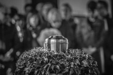 A metal urn with ashes of a dead person on a funeral, with people mourning in the background on a memorial service. Sad grieving moment at the end of a life. Last farewell to a person in an urn.の写真素材