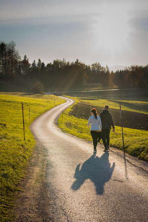 A couple is walking together on an asphalt road towards the sunset on a cold autumn evening with sunset just about to happen. Easy afternoon autumn walk.の写真素材