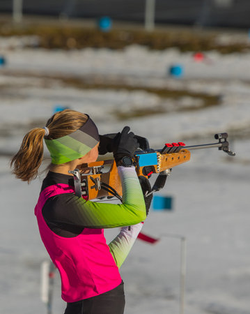 Female biathlon racer is standing on the ground and aiming her rifle. Biathlete woman on a shooting range, firing standing up.の写真素材