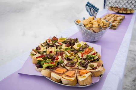 Buffet with sandwiches made of bread, cheese, salami, ham, prosciutto, olives,.. in winter setting on purple table on a ski slope.の写真素材