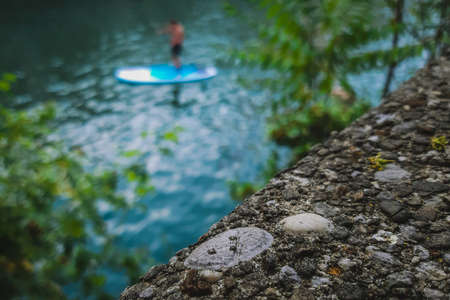 Close up or macro of a stone wall or concrete edge next to a river with visible sup paddler or SUP paddle person standing in blurred background. Good for conceptual photoの写真素材
