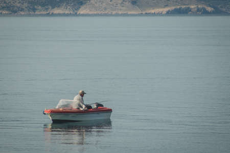 An older fisherman on a small red and white boat on the sea. Fisherman is seen smoking and holding a fishing stick.の写真素材