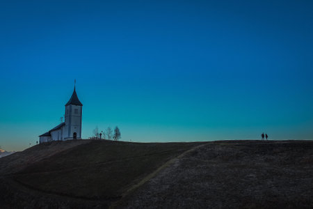 Photo of cute fantastic charming Saint Primoz church on a small hill with mountains in background at sunset, Jamnik village, Slovenia, Europe with picturesque church in early spring and some people as silhouettesの写真素材