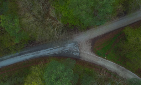 Y shaped crossroads visible from above. Green trees and grey road with transition from asphalt to...