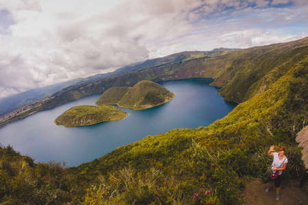 Looking towards the island on famous Lake Cuicocha in Ecuador. Panorama over a blue lake, surrounded with lush greens and white clouds with a young woman waving hi.の写真素材