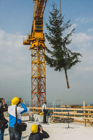 A construction crane is lifting a spruce tree on the top of the building. Photographers are taking photos of the event, as it embarks the end of a construction period on the building.の写真素材