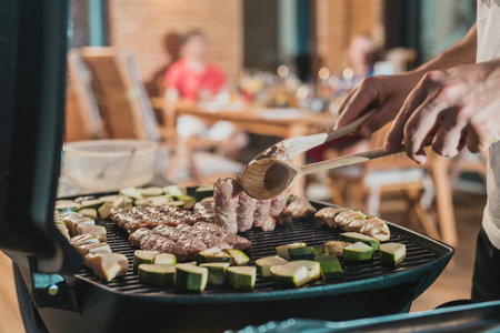 Hands of a man seen flipping cevapcici meal on a grill at an evening party setting. Other guests waiting for their meal in the background.の写真素材