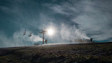 Silhouette of a snow cannon or artificial snow maker on a ski slope on a sunny day. Visible sun flare behind the exhaust of a cannonの写真素材