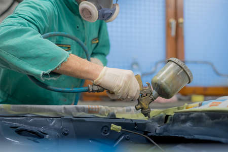 Man in green overalls preparing an engine bay of a car for a final coat of paint. Man spraying grey liquid putty or stucco on the car panels as a last layer before final paint coat.の写真素材