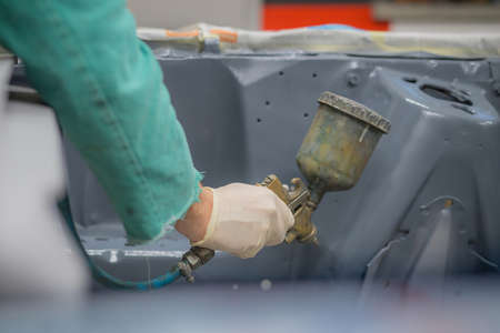 Man in green overalls preparing an engine bay of a car for a final coat of paint. Man spraying grey liquid putty or stucco on the car panels as a last layer before final paint coat.の写真素材