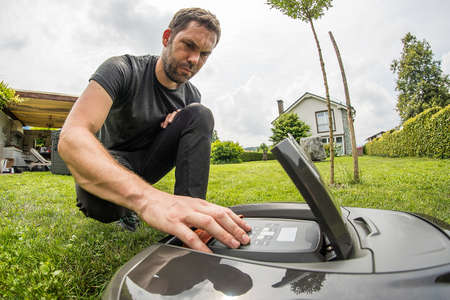 Young caucasian male is setting a robotic mower while kneeling in a nice green garden with blue skies.の写真素材
