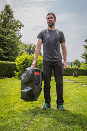 Young caucasian male is holding a robotic mower in his hand while standing in a nice green garden...