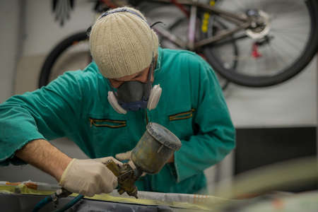 Man in green overalls preparing an engine bay of a car for a final coat of paint. Man spraying grey liquid putty or stucco on the car panels as a last layer before final paint coat.の写真素材