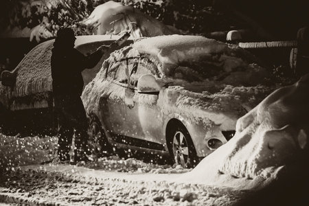 Silhouette of a person cleaning snow and frost a passenger car on a street with the help of a brush in the morning getting ready for the morning commute. Cleaning the car from snow for a commute.の写真素材