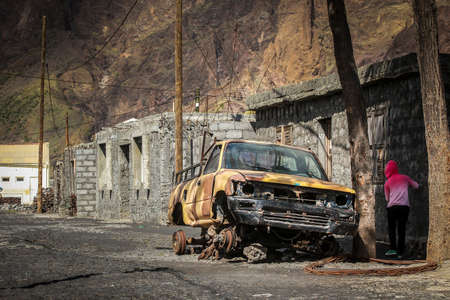 Destroyed and burnt down army pickup truck without wheels on the street on island of Fogo on Cabo Verde islands, with a kid playing around.の写真素材