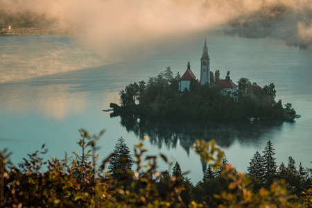 Panoramic photo of lake Bled island with church on a cold hazy foggy early autumn morning from Ojstrica vantage point. Visible fog coming out of water.の写真素材