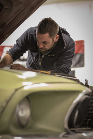 A young man is reaching under the hood of an old vintage car fixing the engine. Man struggling to...
