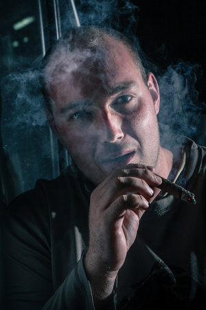 A young man with a serious expression is smoking a cigar in a dark environment, with a cloud of white smoke seen exhaling. Noir photo of a man smoking a cigar.の写真素材