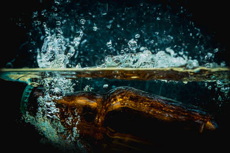 Brown beer bottle falls into water making big splash of drops and spray on a dark background. Juicy and fresh concept of refreshing drink in water.の写真素材